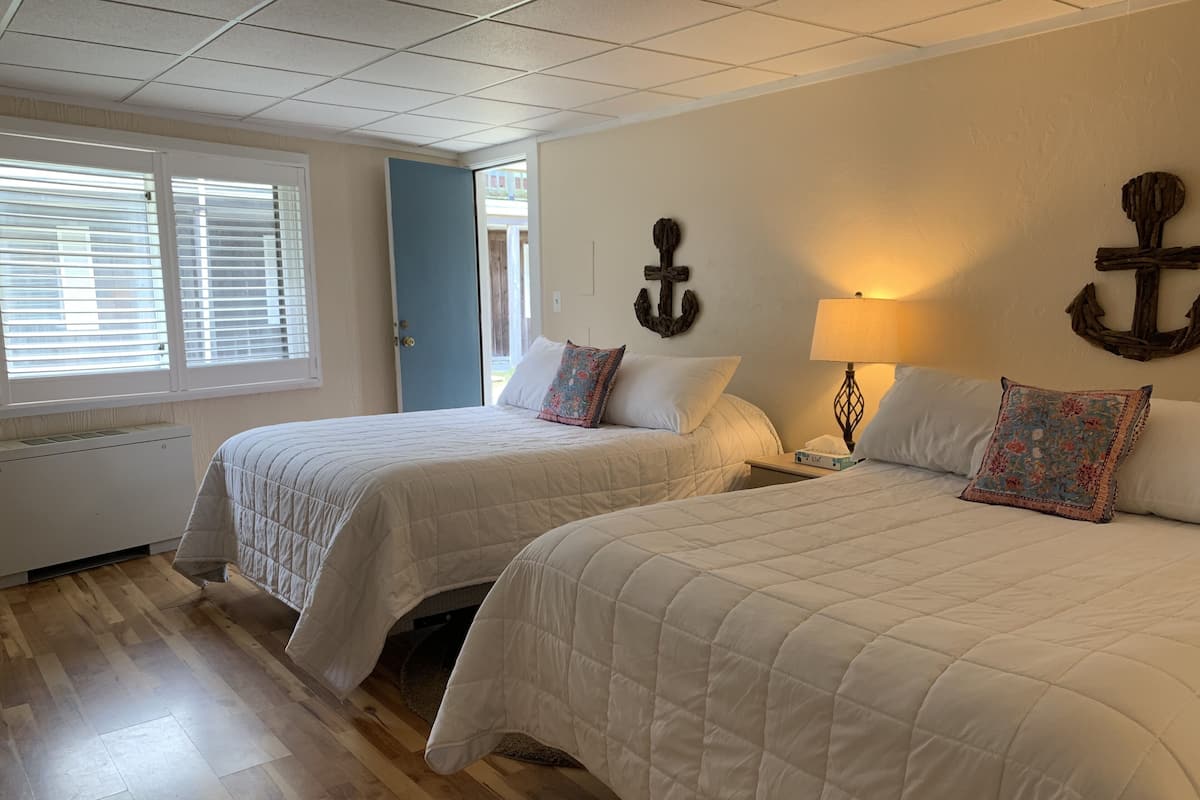 A hotel room with two beds, a wooden floor, a window with blinds, and a wall-mounted lamp.