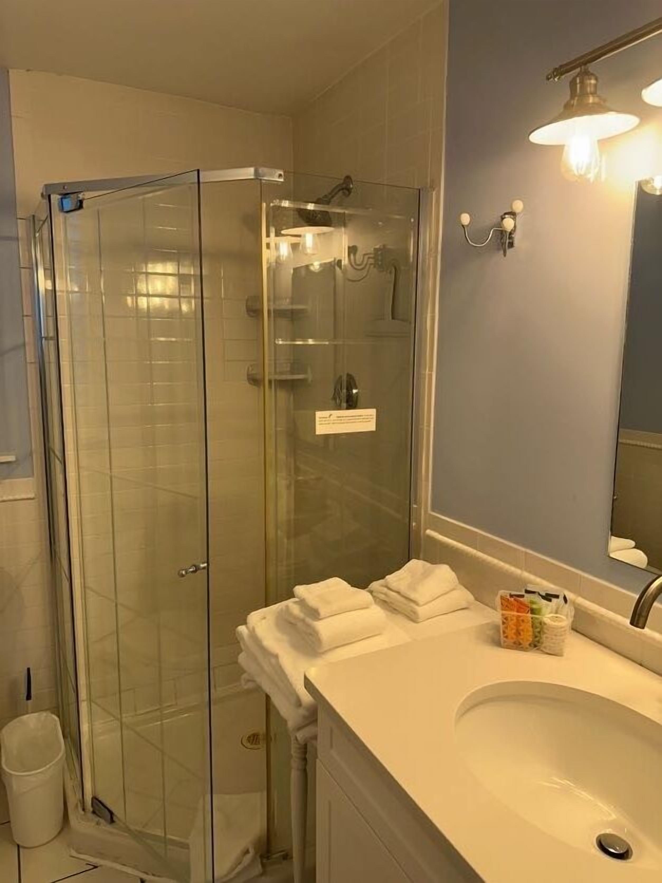 Un baño con ducha envuelta en vidrio, un lavabo con espejo y un cubo de basura.