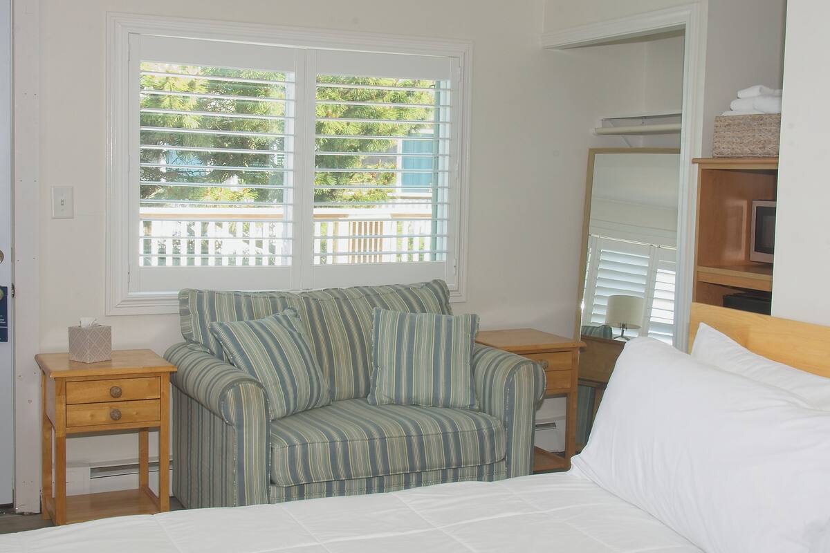A bedroom with a bed, a striped sofa, a wooden nightstand, a bedside table, and a window with blinds.