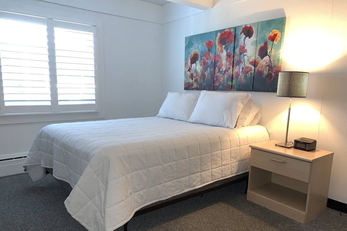 A neatly made bed with white bedding, a bedside lamp, and a painting of flowers on the wall.