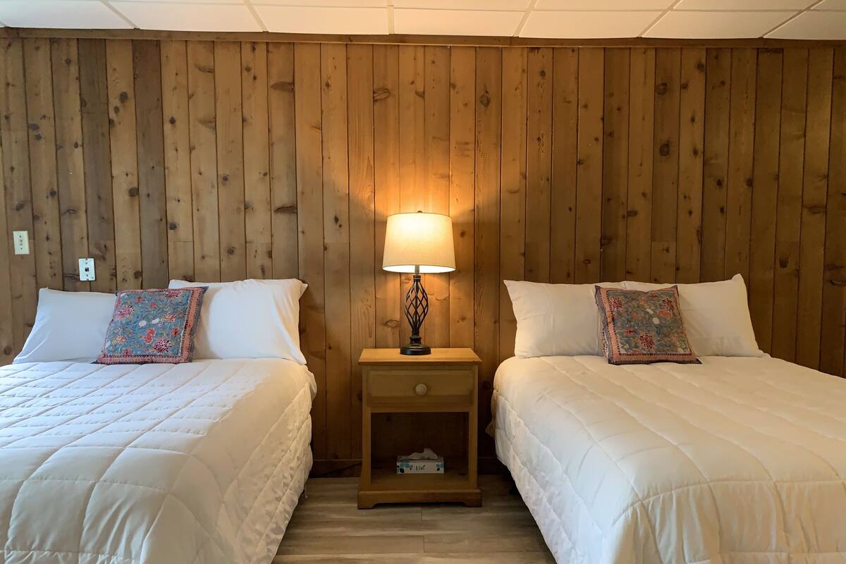 A room with two beds, each with a white duvet and a floral pillow, flanked by wooden walls. A nightstand with a lamp is positioned between the beds.