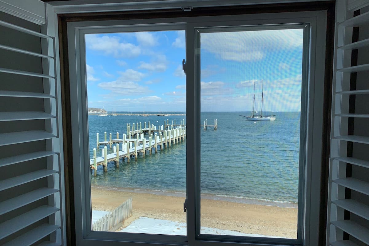 A view through a window with white blinds, overlooking a beach with a wooden pier extending into calm waters, and a sailboat anchored in the distance.