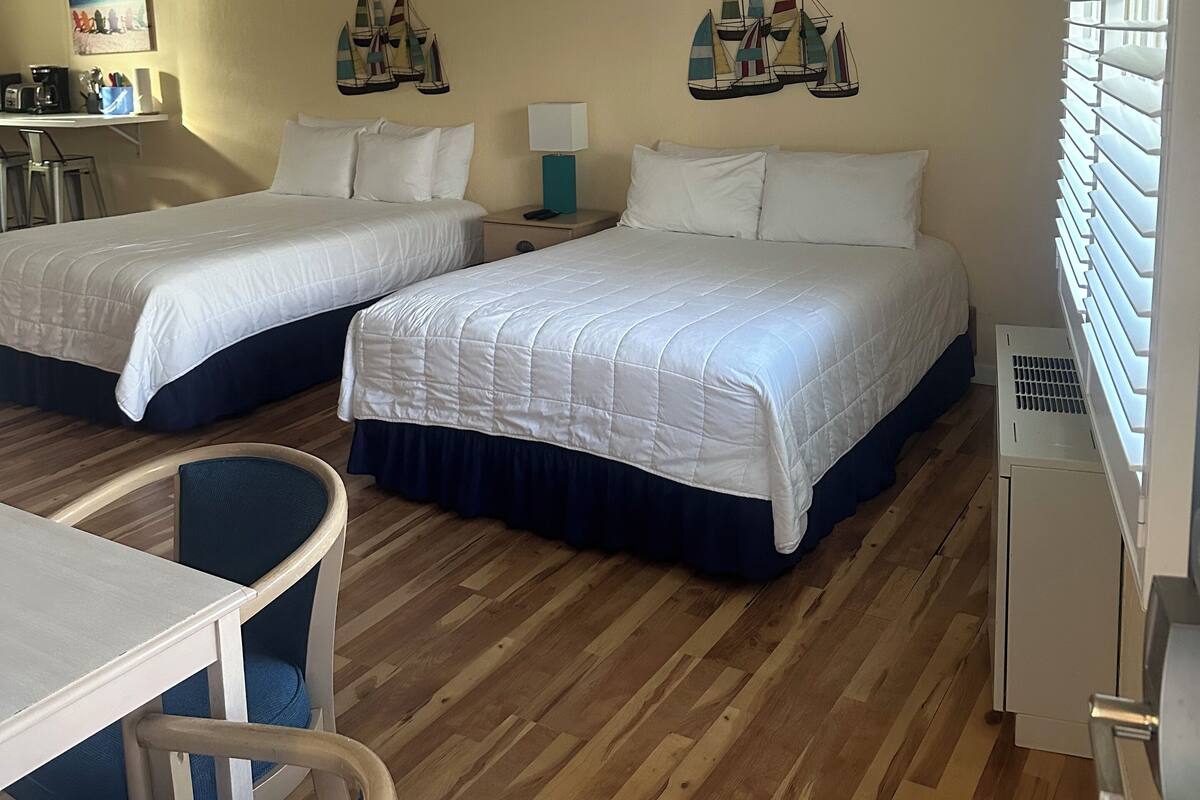 A hotel room with two beds, wooden flooring, a desk with chairs, and a window with blinds.