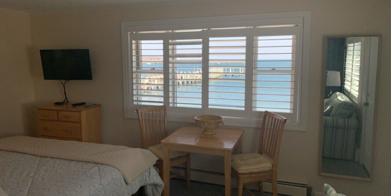 A room with a bed, a TV on a wooden dresser, a table with chairs, and a window with a view of the sea and a pier.