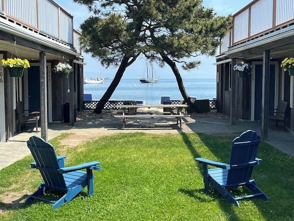 A patio area with two Adirondack chairs facing a view of the sea, a picnic table, and a tree.