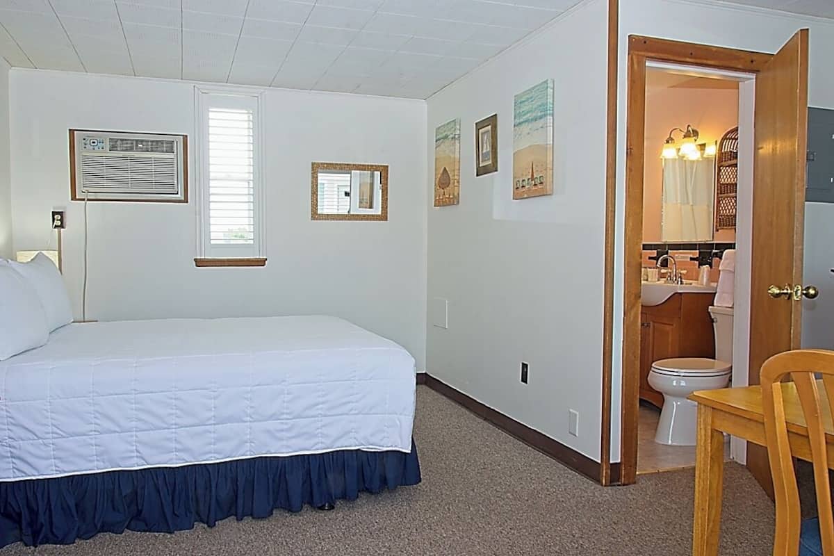 A bedroom with a bed, a window, a wall-mounted air conditioner, and a bathroom visible through an open door.