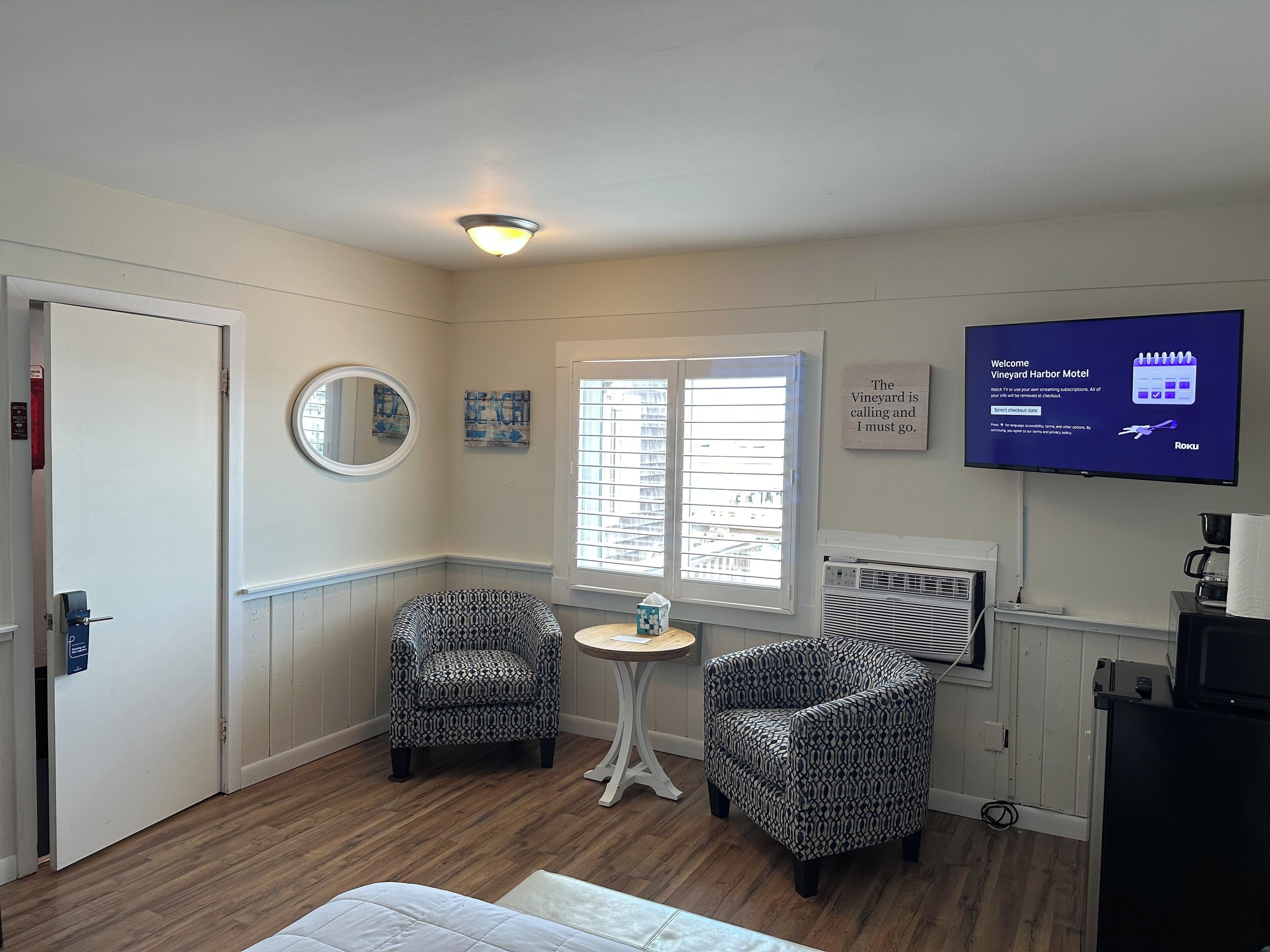 A hotel room with a wooden floor, two patterned armchairs, a small round table, a wall-mounted TV, and a window with blinds.