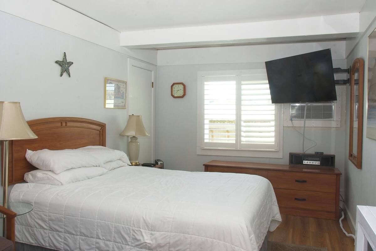 A bedroom with a bed, bedside lamps, a dresser, a TV, a clock, and a window with blinds.