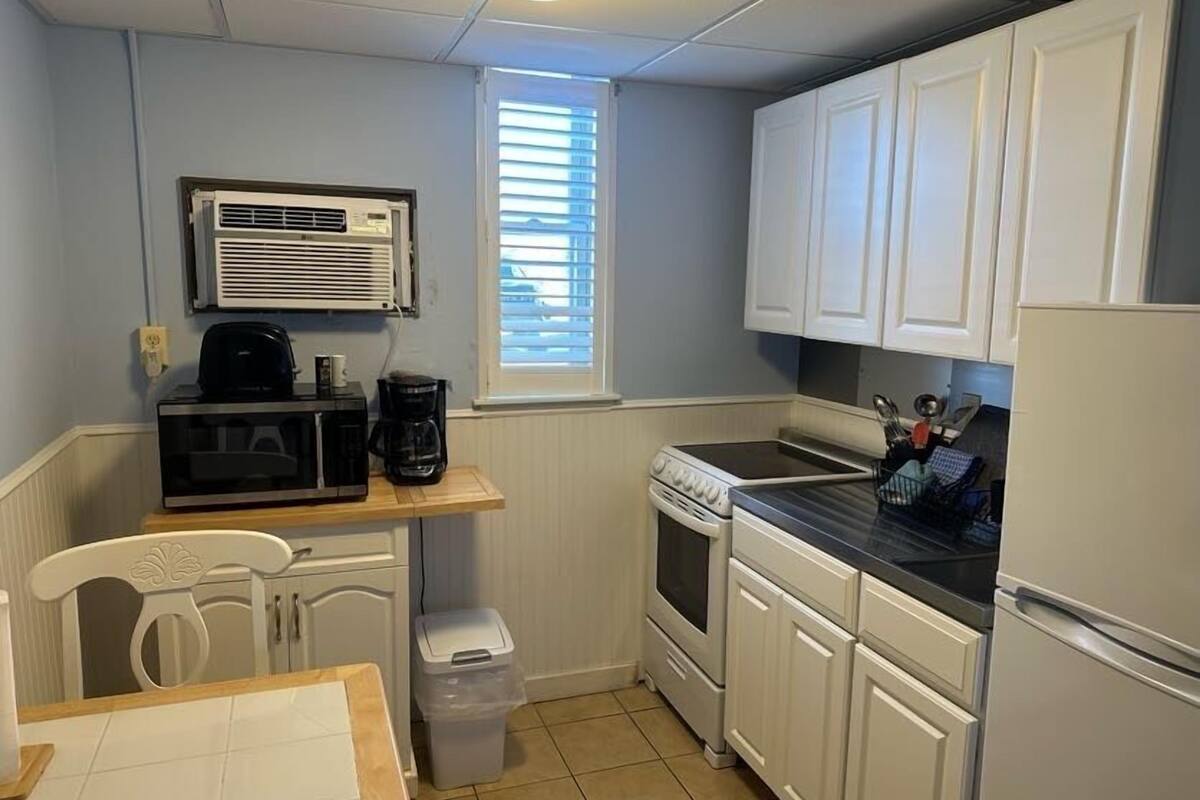 A compact kitchen with white cabinets, a stove, microwave, and a small dining area with a chair and table.