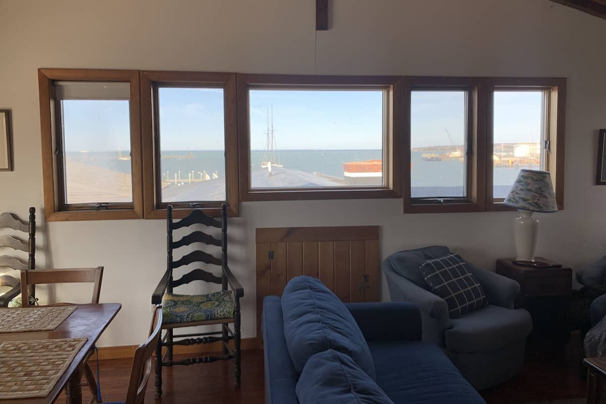 A living room with a view of the sea through large windows, a blue sofa, a wooden chair, and a lamp on a side table.