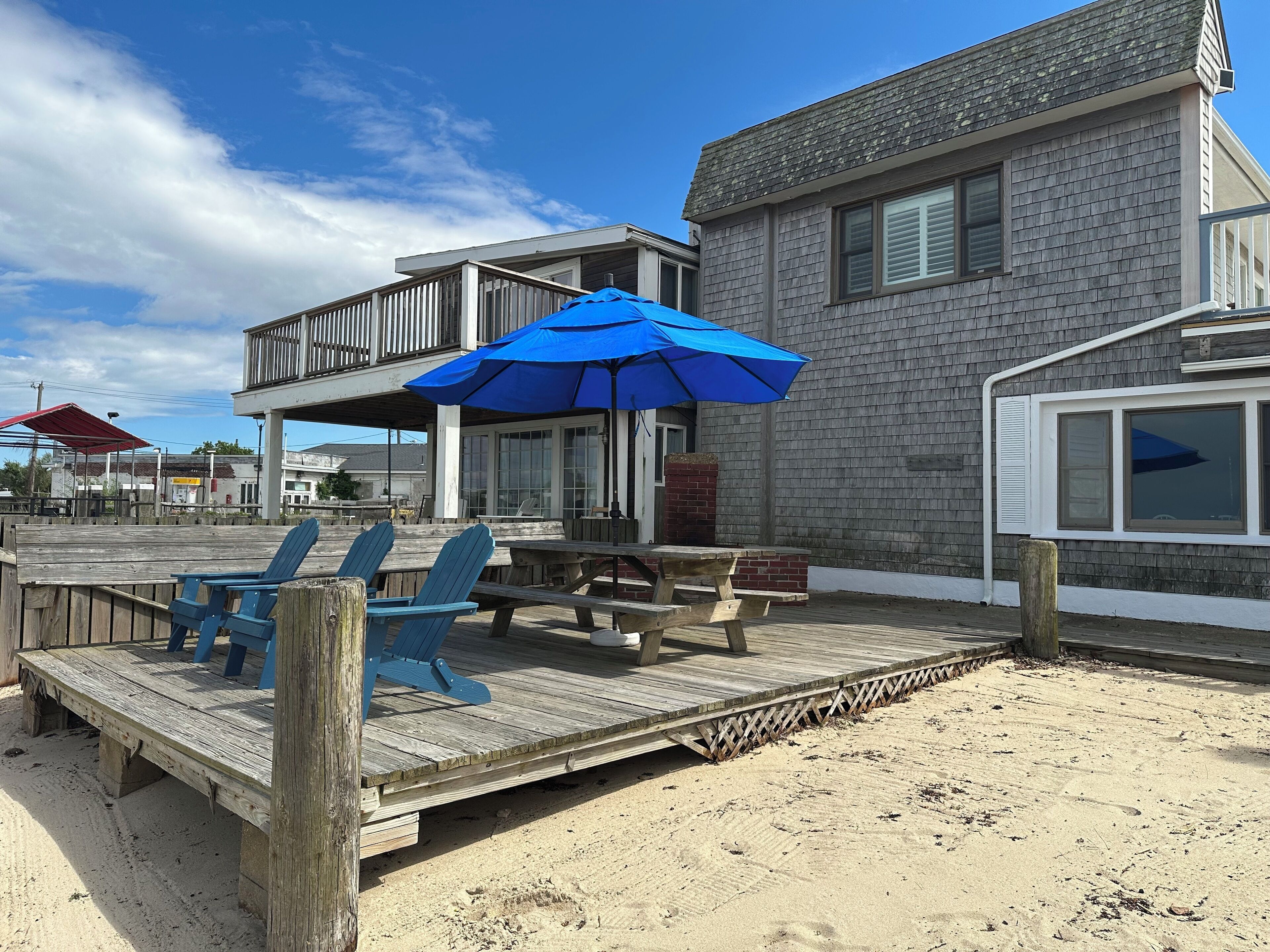 Una terraza de madera frente a la playa, con sillas azules y una mesa de picnic, una sombrilla azul, y un edificio con balcón y contraventanas.