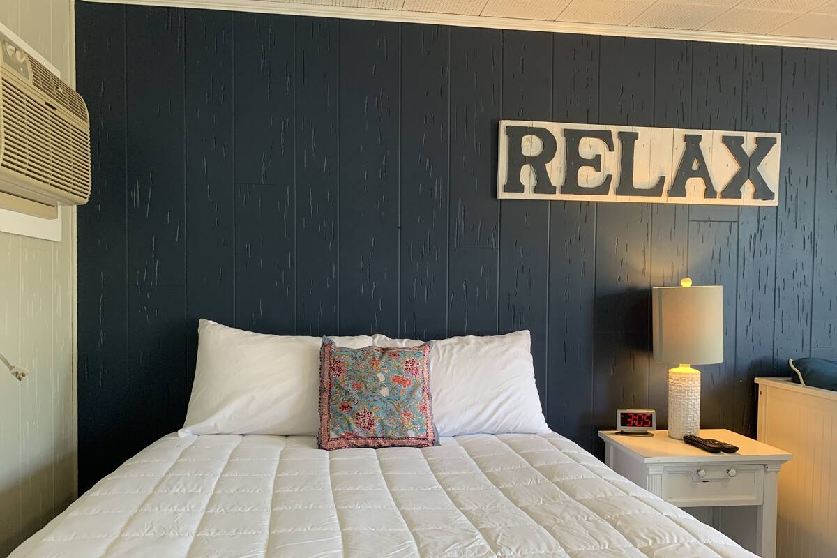 A neatly made bed with a quilted white duvet, a decorative pillow, and a wall-mounted sign that reads "RELAX".