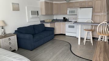 Une cuisine compacte équipée d’un four à micro-ondes, d’une cuisinière et d’un réfrigérateur blanc. Un canapé bleu et une table à manger en bois, accompagnés de deux chaises, se trouvent dans l’espace salon.