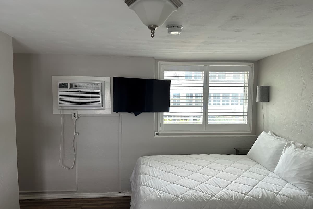 A bedroom with a bed, an air conditioning unit, a TV, and a window with blinds.