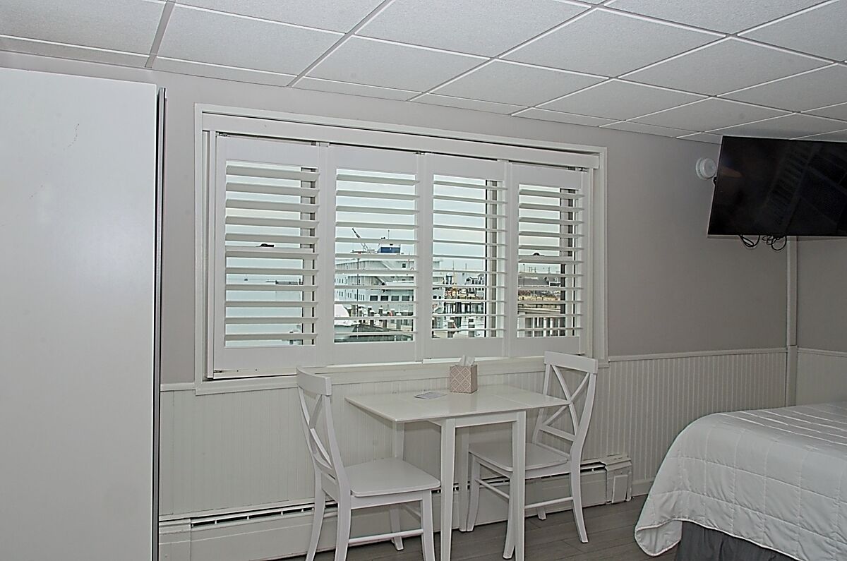 Een kamer met een bed, een aan de muur bevestigde televisie, een raam met zonwering en een tafel met twee stoelen.