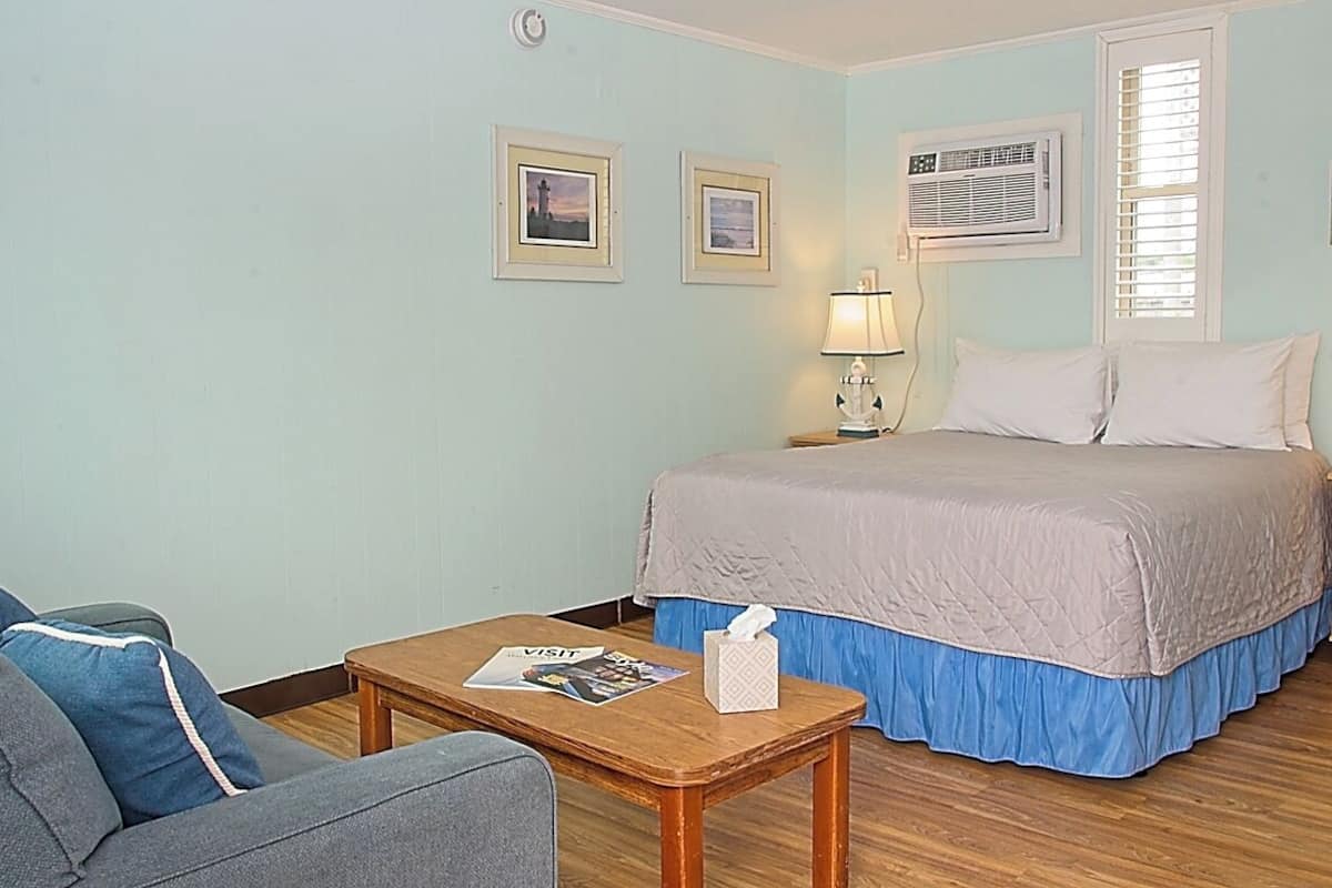 A hotel room with a bed, a nightstand with a lamp, a table with a magazine and tissues, and two framed pictures on the wall.