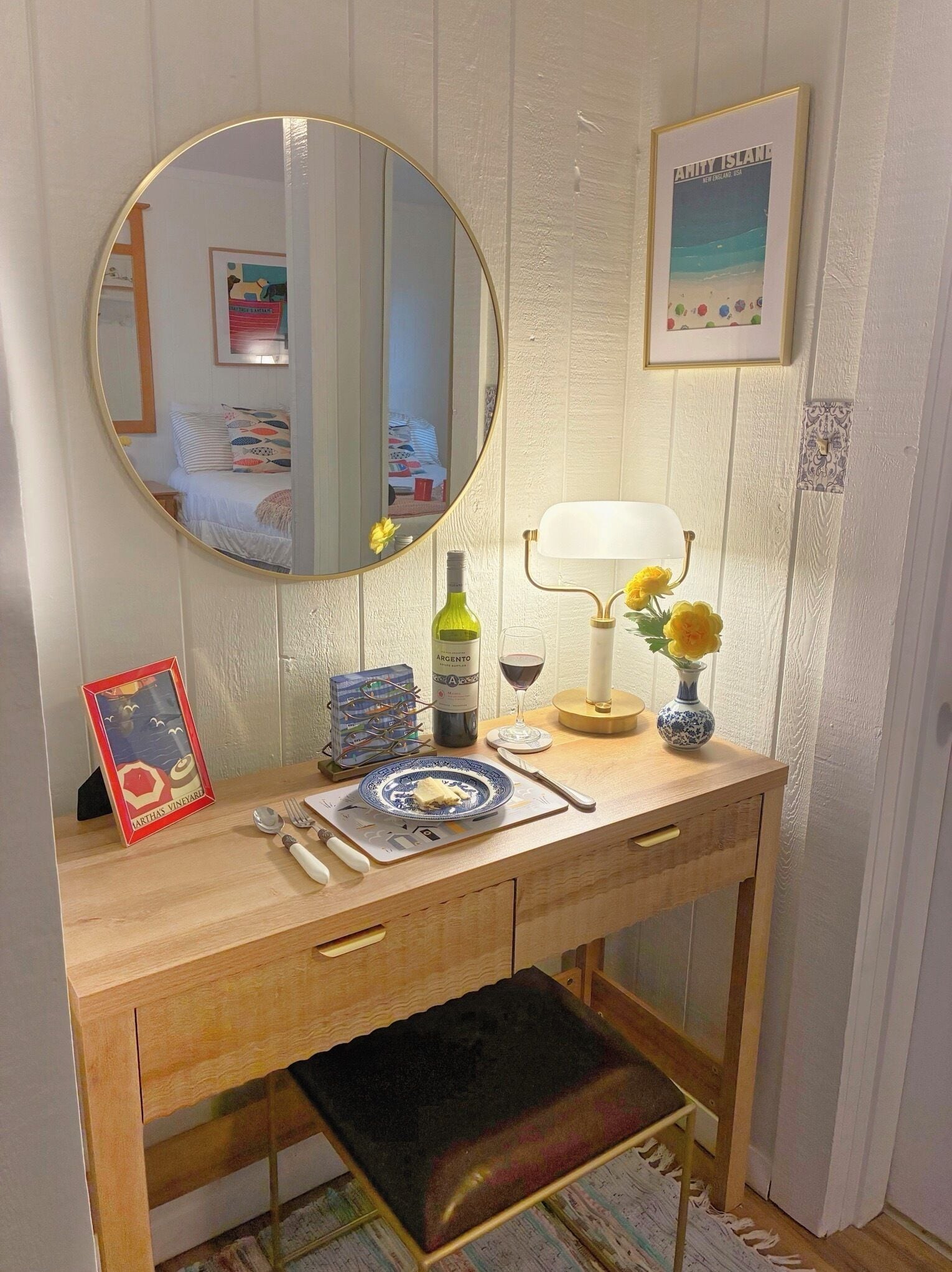 A wooden desk with a mirror, a framed picture, a wine bottle, a candle, and a vase with flowers.