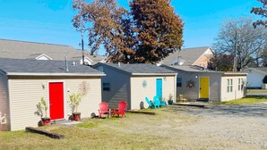 Fila de casas móviles de un solo piso con puertas de colores llamativos, un camino de grava y un área ajardinada con plantas en macetas y mobiliario exterior.