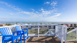 Une terrasse en bois avec des chaises bleues, une balustrade blanche et une vue sur l’océan et la plage.