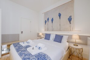 Una camera d'albergo con un letto grande, un comodino con una lampada, un'opera d'arte appesa al muro raffigurante uccelli e una porta.