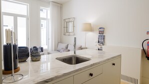 Een moderne keuken met een marmeren aanrecht, een roestvrijstalen spoelbak en een inbouwoven. Er is een raam met witte gordijnen, een vloerlamp en een klein plankje met decoratieve objecten.