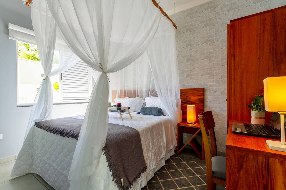 Quarto com cama, mesa de cabeceira, cadeira e escrivaninha com um laptop.
