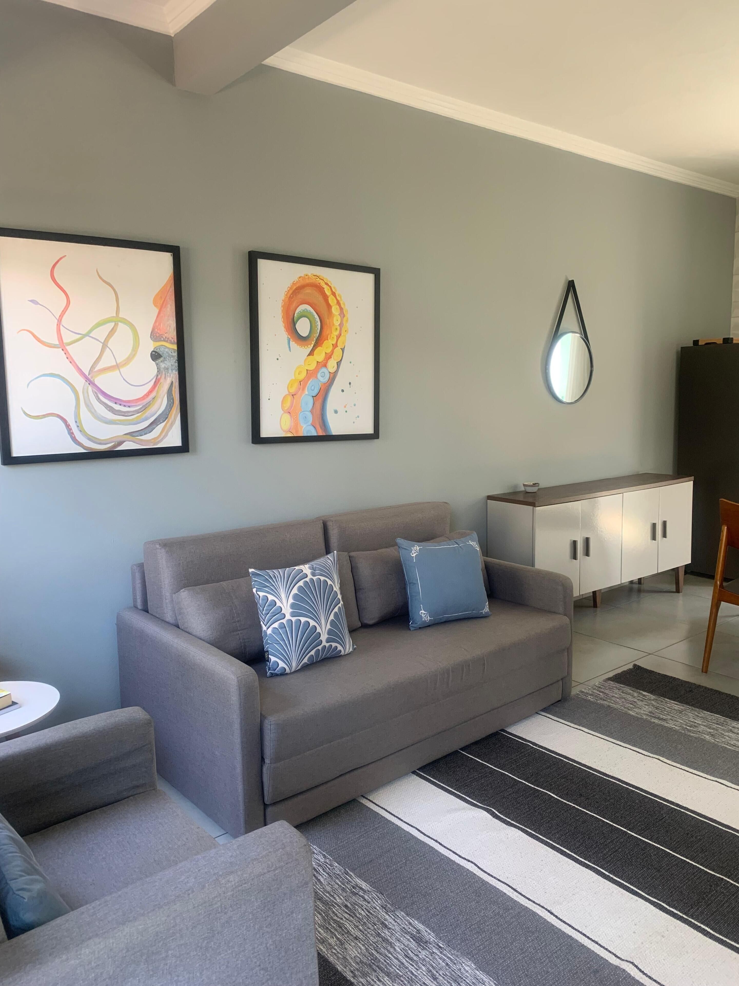 Un salón moderno con un sofá gris, dos cojines estampados, un espejo redondo y decoración abstracta en la pared.