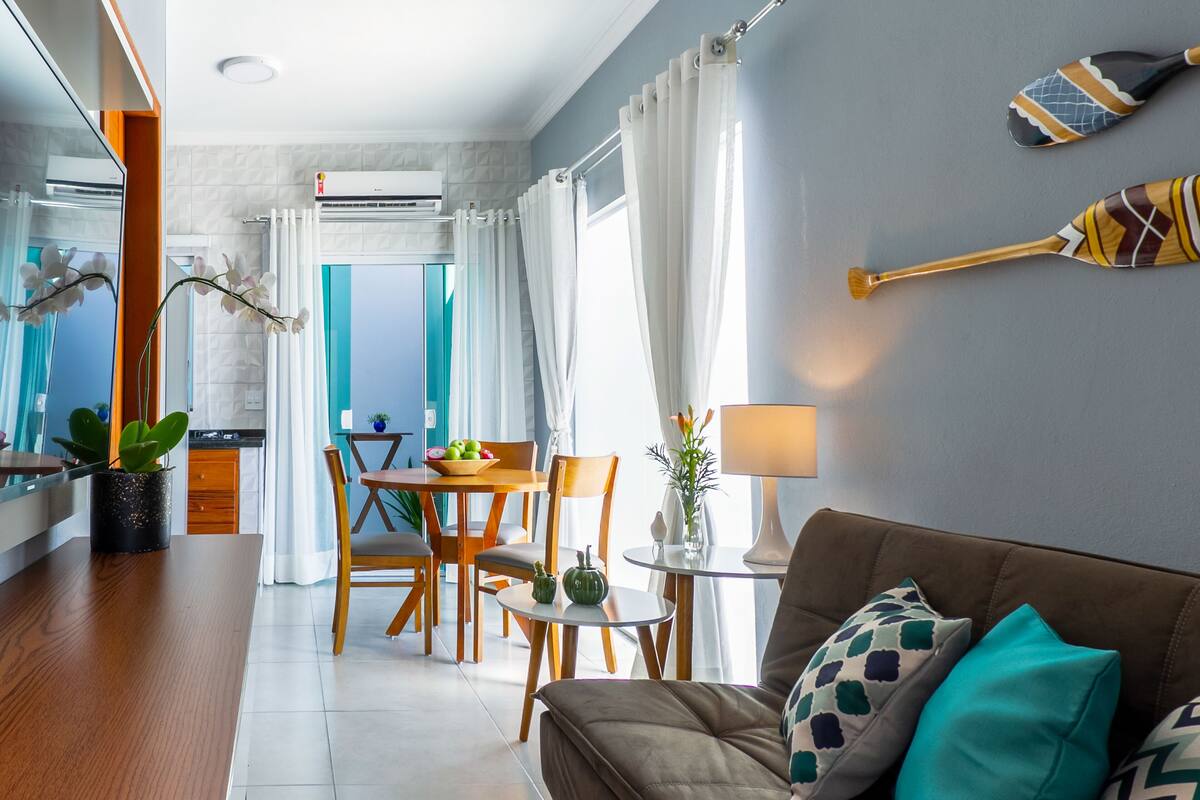 Uma sala de estar moderna com um sofá marrom, almofadas verde-água, uma área de jantar com móveis de madeira e uma cozinha visível ao fundo.