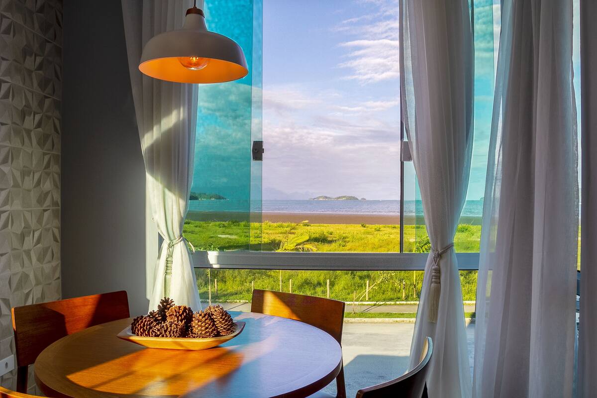 Uma área de refeições com mesa, cadeiras, uma tigela de pinhas e vista para uma praia através de cortinas translúcidas.