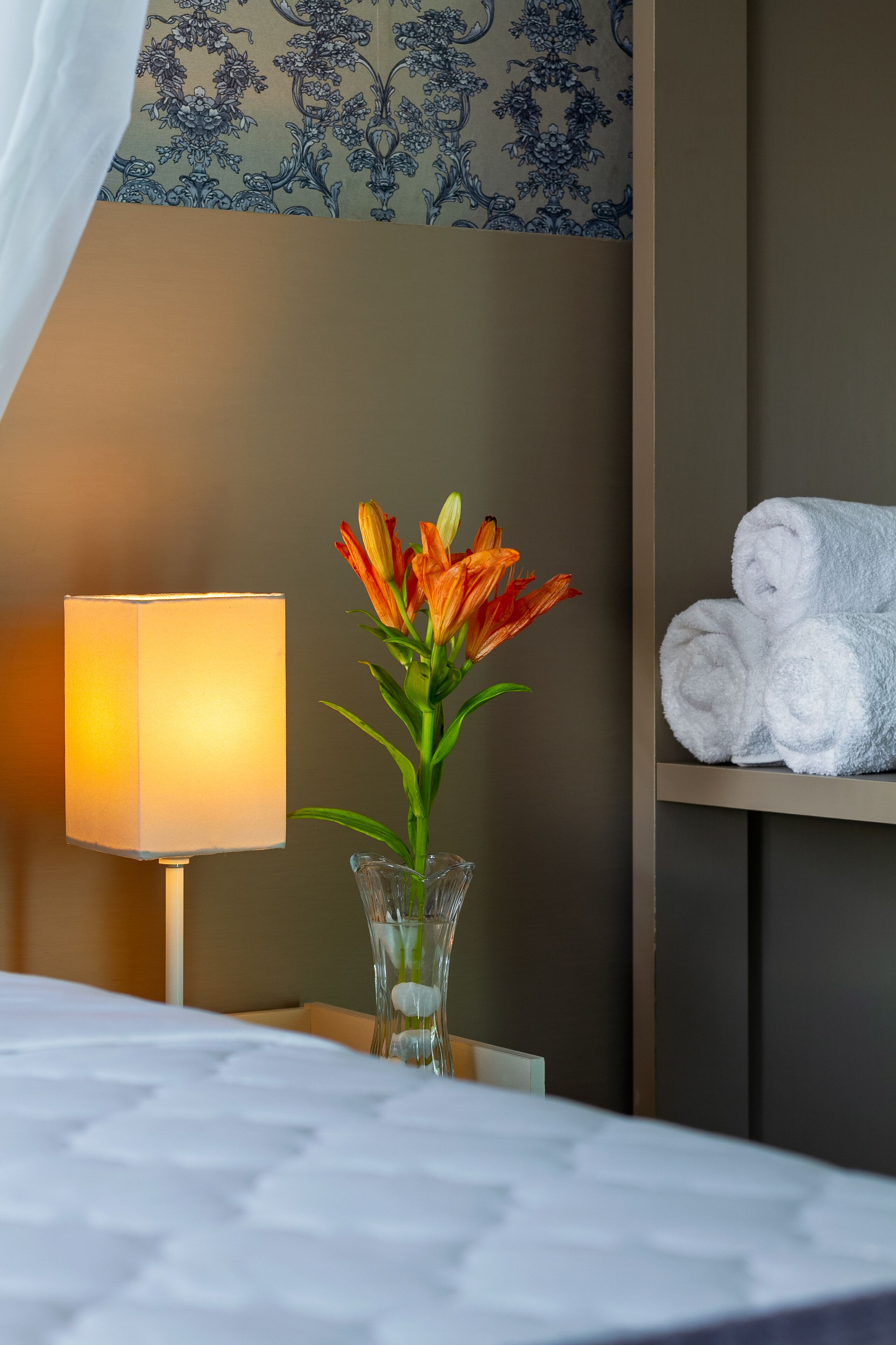 Habitació d'hotel amb un llit, una làmpada, un vas amb flors i una estant amb tovalloles.