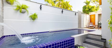 A modern outdoor swimming pool with blue tiles, a water feature, and steps leading to a wooden door.