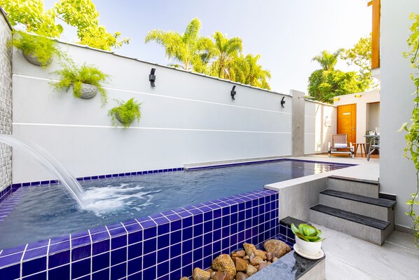 Una piscina moderna al aire libre con azulejos azules, un elemento acuático y escalones que conducen a una puerta de madera.