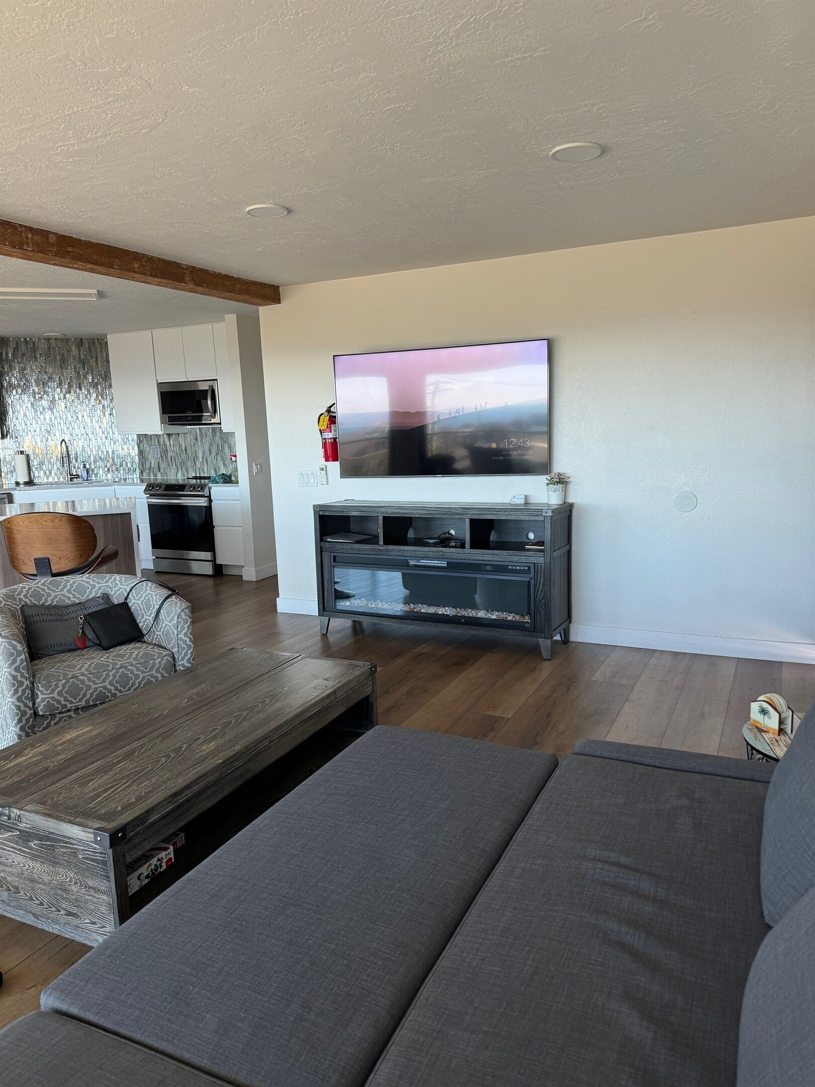A modern living room with a large flat-screen TV mounted on the wall, a grey sofa, a wooden coffee table, and a patterned armchair.