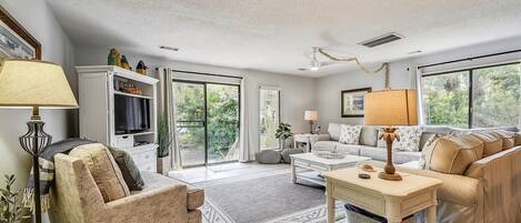 A spacious living room with a sofa, armchair, and a coffee table. There is a television on a cabinet, and a large window providing a view of the outdoors.
