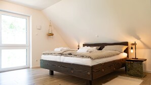 A bedroom with a wooden bed, a bedside table with a lamp, and a view of the outdoors through a large window. - Vacation home Heuliebe, barbecue hut, whirlpool, outdoor sauna (Herbstein)