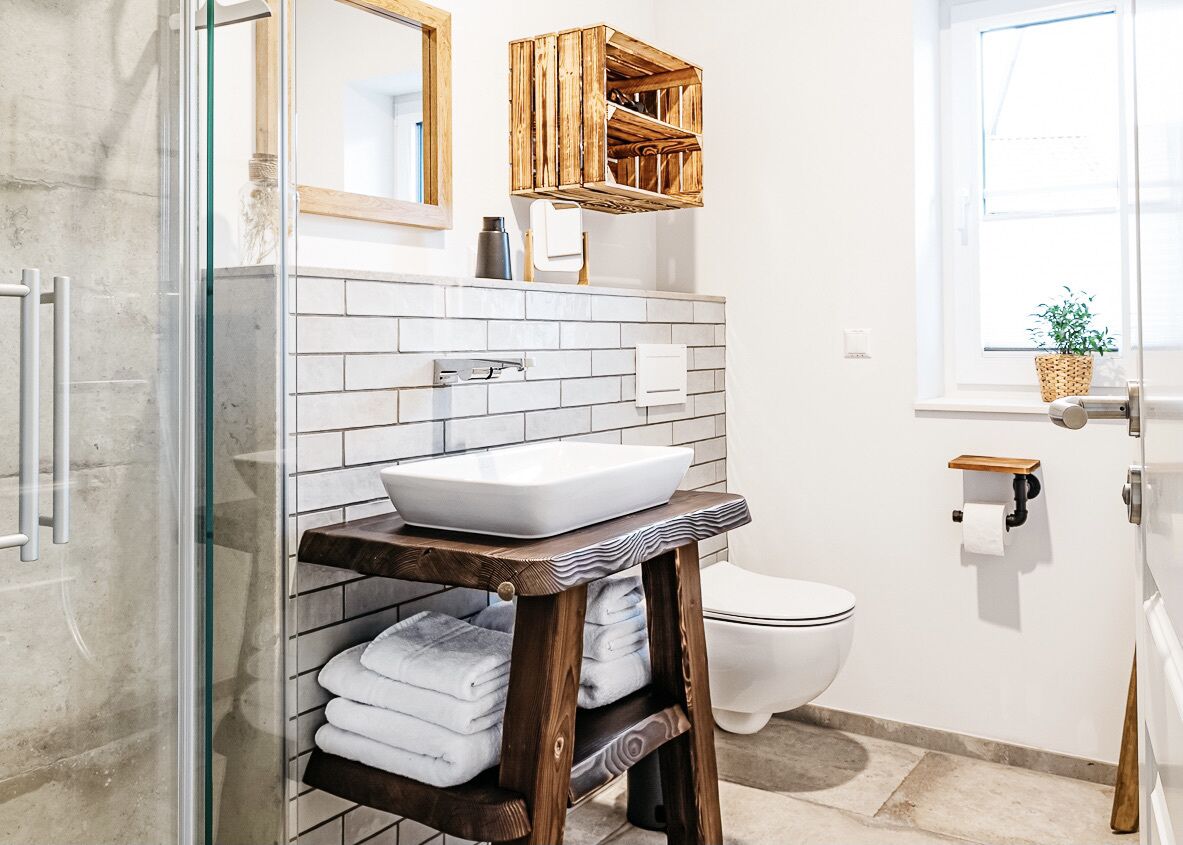 A modern bathroom with a glass shower enclosure, a white sink with a wooden shelf, a white toilet, and a window with a plant.