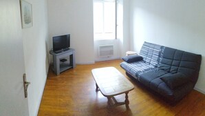 En stue med sofa, et lille bord, et fjernsyn på et stativ og et vindue med persienner.