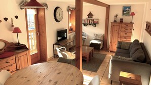 Un accogliente soggiorno con un tavolo da pranzo in legno, un divano e una televisione. Si nota un orologio a parete e una finestra con tende.