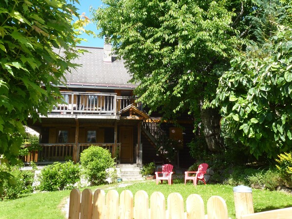 A two-story wooden house with a balcony, surrounded by lush greenery and a wooden fence.