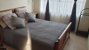 En seng med grå madras og mønstrede puder, et natbord med en lampe, og et vindue med lette gardiner.