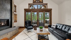 A modern living room with a fireplace, leather furniture, and a large window overlooking a forest.