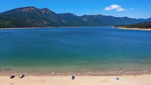 A beach with clear water, mountains in the background, and a few cars on the sand.