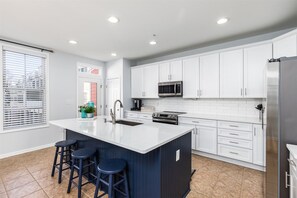 A modern kitchen with white cabinets, a central island with stools, a stainless steel refrigerator, and a window with blinds. - Sunset Island 20 Beach Side Drive (Ocean City)