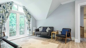Una sala de estar moderna con un sofá gris, una silla de madera, una mesa de centro de vidrio y un amplio ventanal con cortinas.