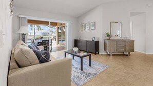 A spacious living room with a sofa, coffee table, and a dining area with a table and chairs. There is a sliding glass door leading to a patio with a pool.