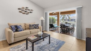 A living room with a beige sofa, patterned cushions, a coffee table with a magazine, a floor lamp, and a view of an outdoor patio with a pool and dining area.