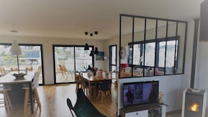 Un vaste espace de vie comprenant une table à manger, des chaises, une cheminée et de grandes fenêtres donnant sur l’extérieur.