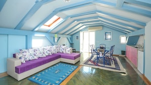 Een ruime zolderkamer met een schuine kap, een paarse bank, een eettafel met stoelen en een keukenhoek.