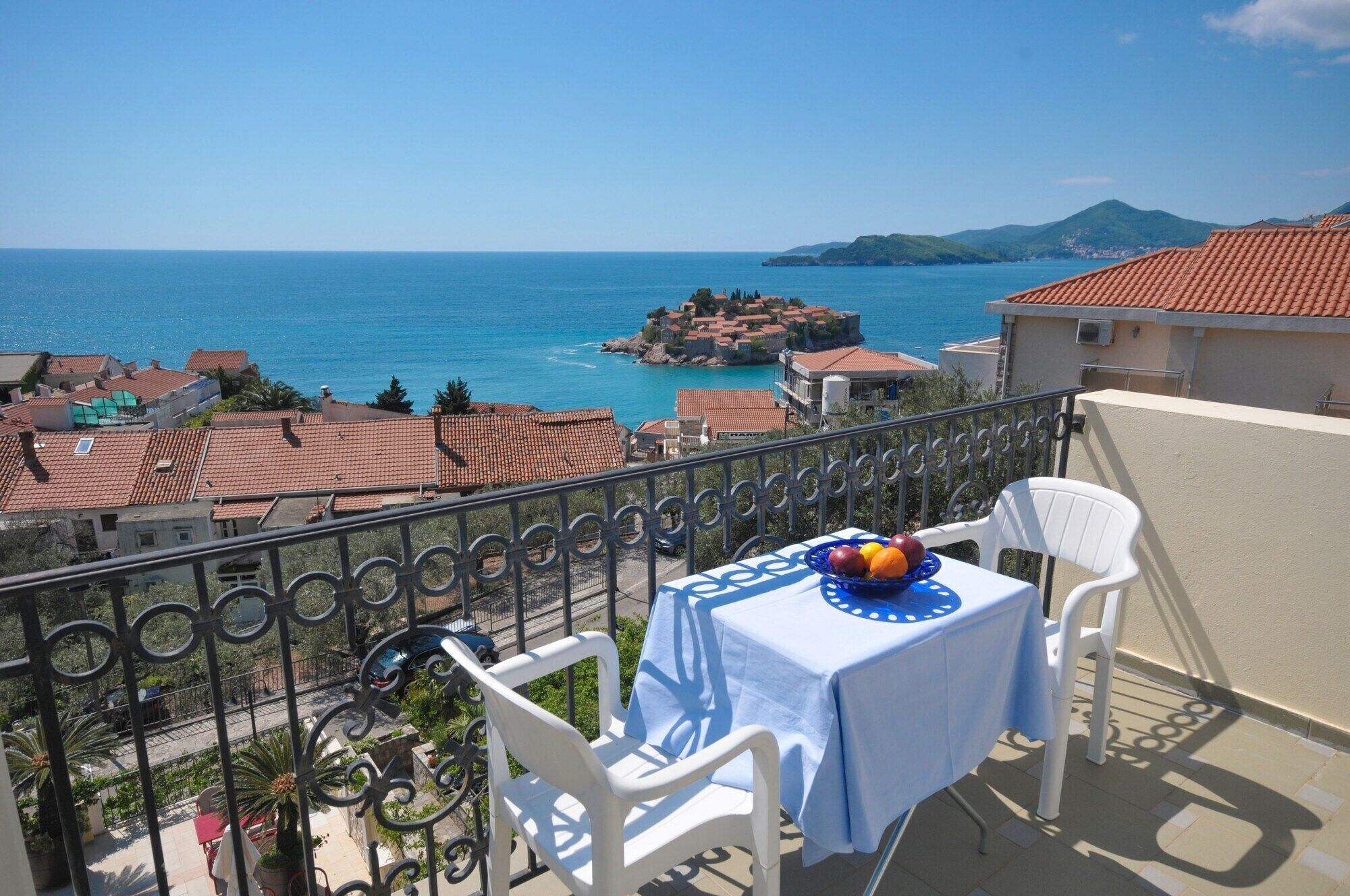 A balcony with a table set for two, overlooking a coastal town with terracotta roofs and a view of the sea.