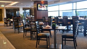 Ein Hotelrestaurant mit mehreren Tischen und Stühlen, einem an einer Säule befestigten Fernseher und großen Fenstern mit Blick ins Freie.